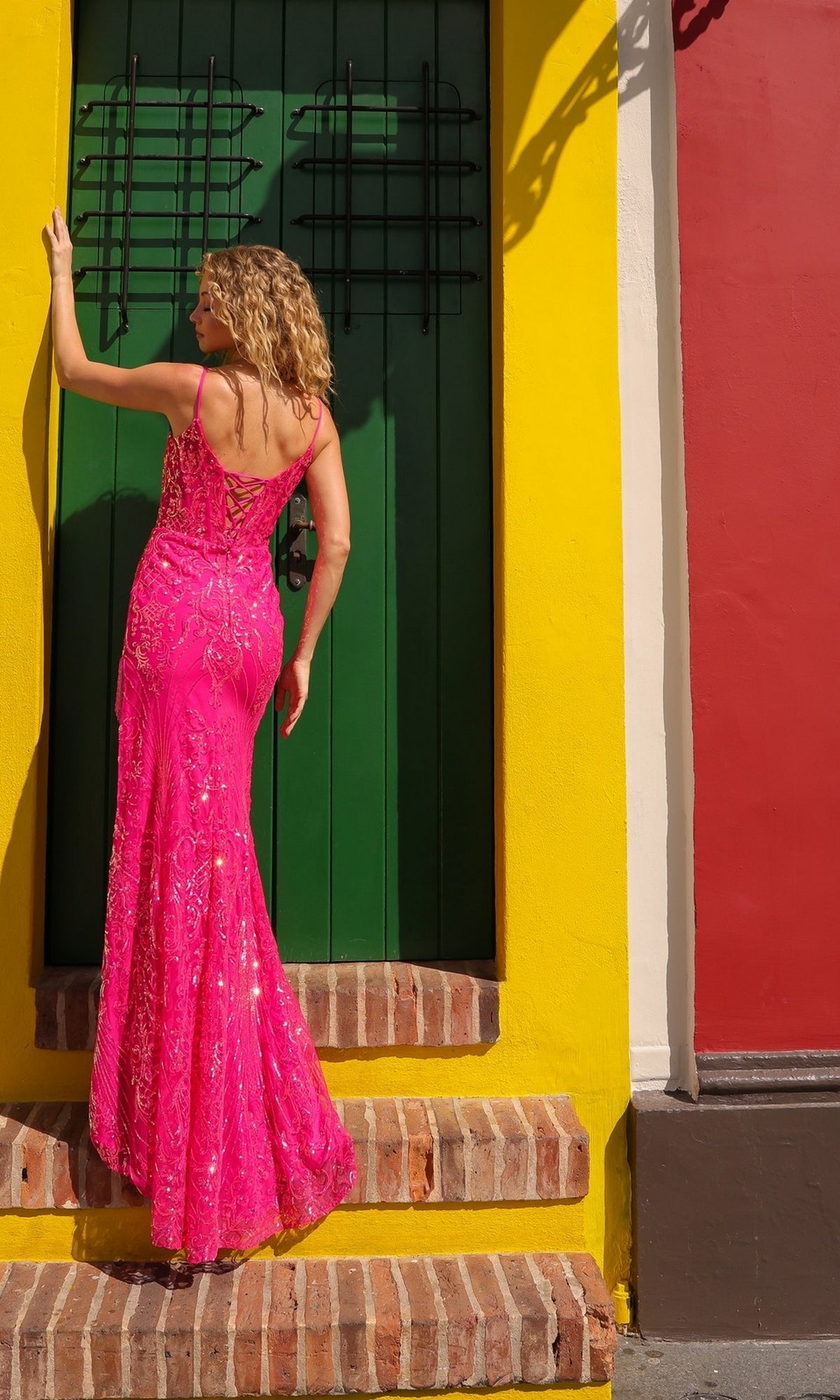 A woman in the Nox Anabel D1465 Fringed Long Sequin Prom Dress poses on brick steps before a green door set in a yellow wall with a red section to the right.